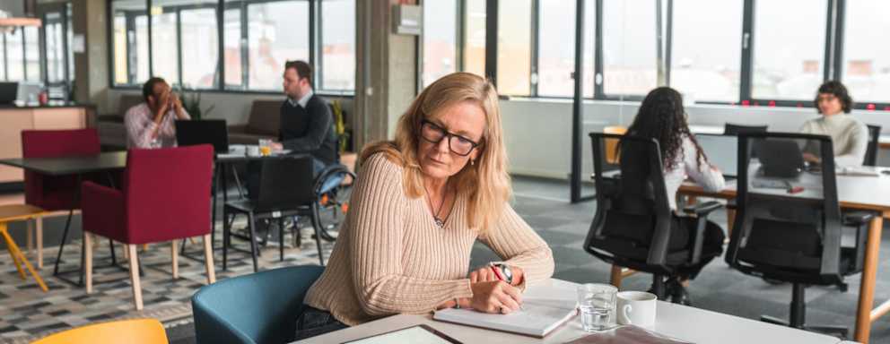 A Caucasian female college student in her late 40s sitting in a common area in a college campus building. She is writing notes on a paper pad to study for an upcoming exam. She has a digital tablet, a cup, a glass and notebooks scattered around on the table top. Other women and men are studying in the background.