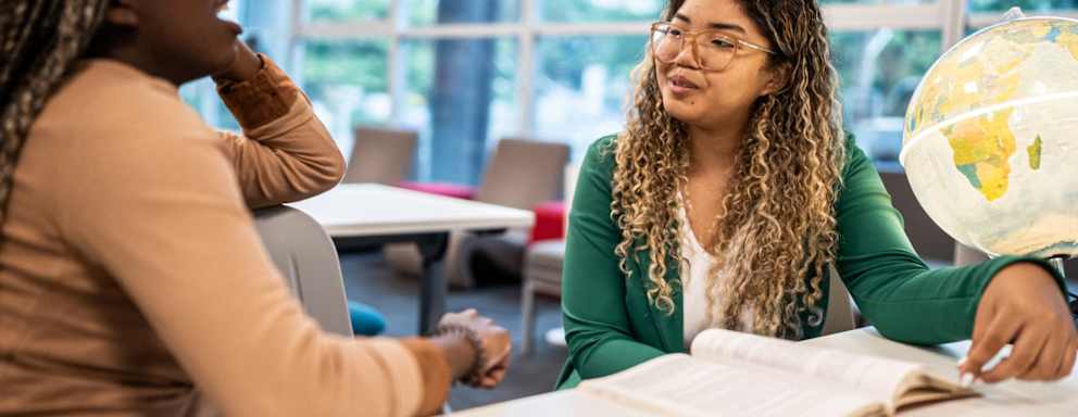 A pair of female college students sitting and talking at a table in a campus library.