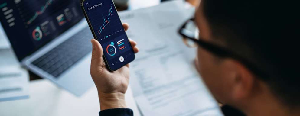 Young Asian man managing finance and investment online, analyzing stock market trades with smartphone and laptop.