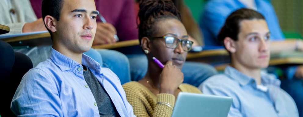 A diverse group of college students are sitting in a university classroom. They are taking notes while listening to their professor (located offscreen) during the lecture.