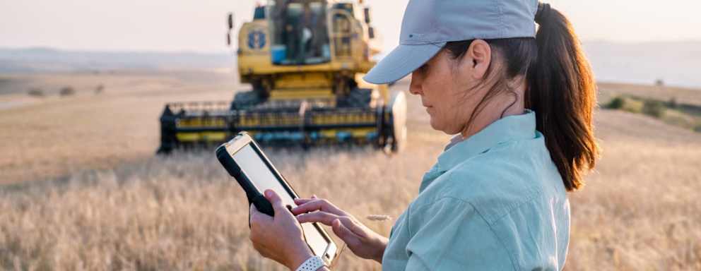 Caucasian female agribusiness professional wearing causal clothes and a baseball cap stands in a grain field on a farm. She is holding a digital tablet in her left hand to write down notes and data.