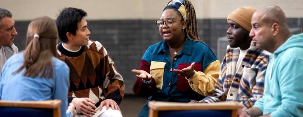 A Black female social worker leading the conversation at a support group meeting at a community center. She is sitting with a diverse group of adult men and women in a circle, who are discussing about mental health. They are all wearing casual clothing.