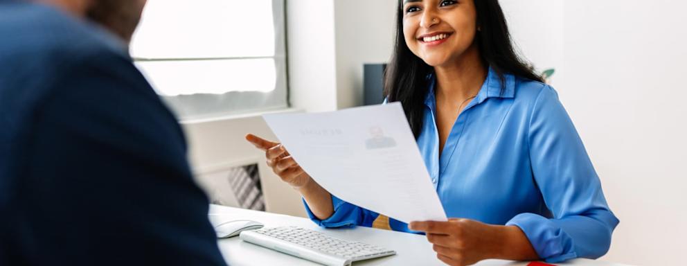 Indian female HR recruitment manager holding a paper resume in her hands while having an interview in a modern office. She is looking and listening to a Caucasian male job candidate, pictured offscreen.