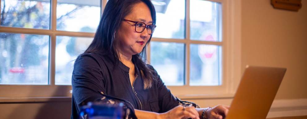 Asian American female business student in her mid-40s typing an assignment for her online class at home. She is sitting at her dining room table, and is wearing glasses and business casual clothes.