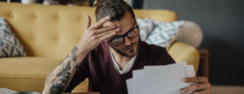 Caucasian male college student sitting at his living room coffee table. He has a worried look on his face while he's holding several paper bills in his hand.