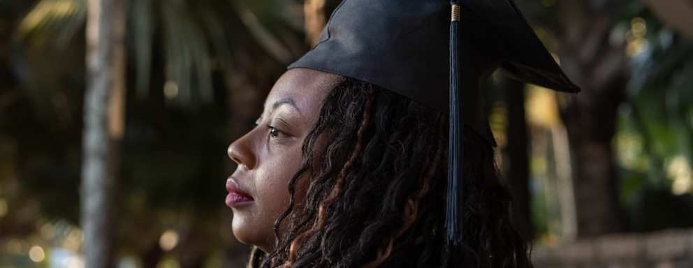 Adult Black female woman dressed in a black graduation cap and gown at her college graduation ceremony.