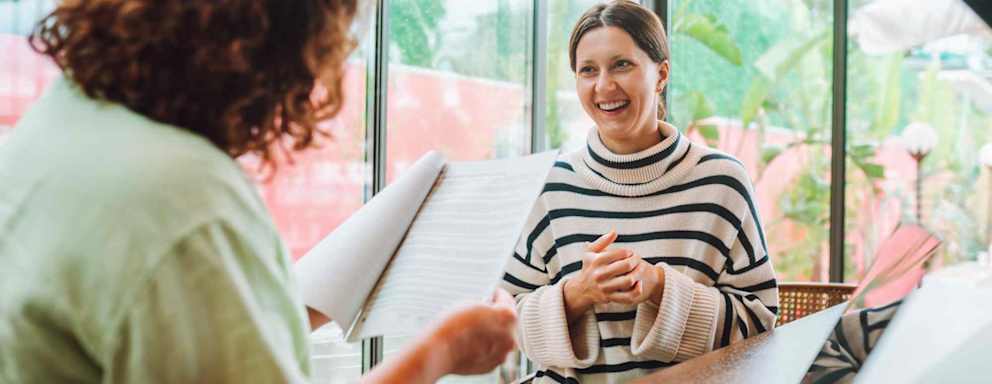Caucasian female hiring manager meeting with a Caucasian female job candidate during an onsite interview. The hiring manager is looking over a paper copy of the candidate's resume.