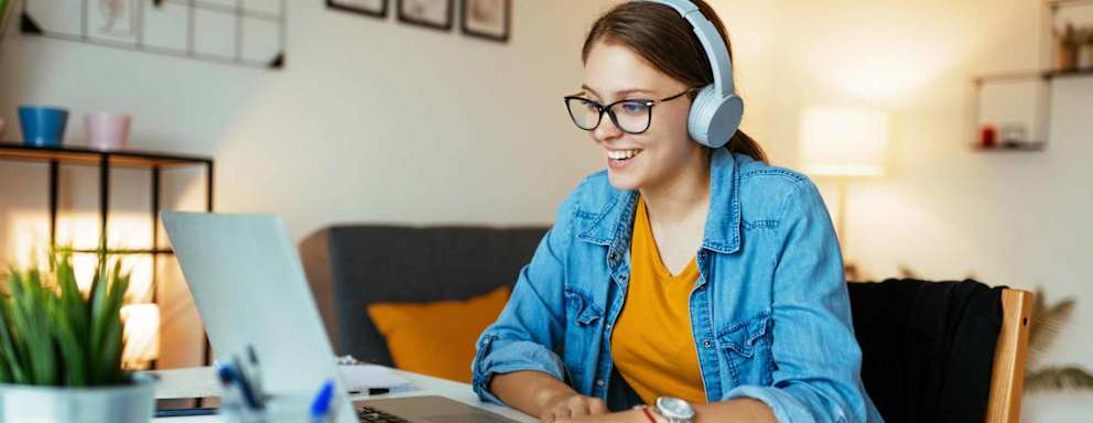 A Caucasian female college student sitting at a table in her living room at home. She is wearing glasses and casual clothes. She has her laptop open and is smiling and chatting with other students before the start of an online class.