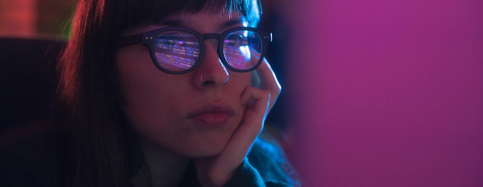 Caucasian female programmer wearing glasses is coding on her desktop computer. She is resting her head on her hand.