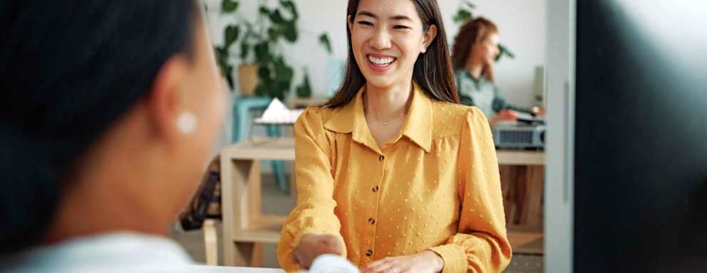 Two female business professionals are shaking hands and smiling in a corporate office. One is Asian American and the other is Black.