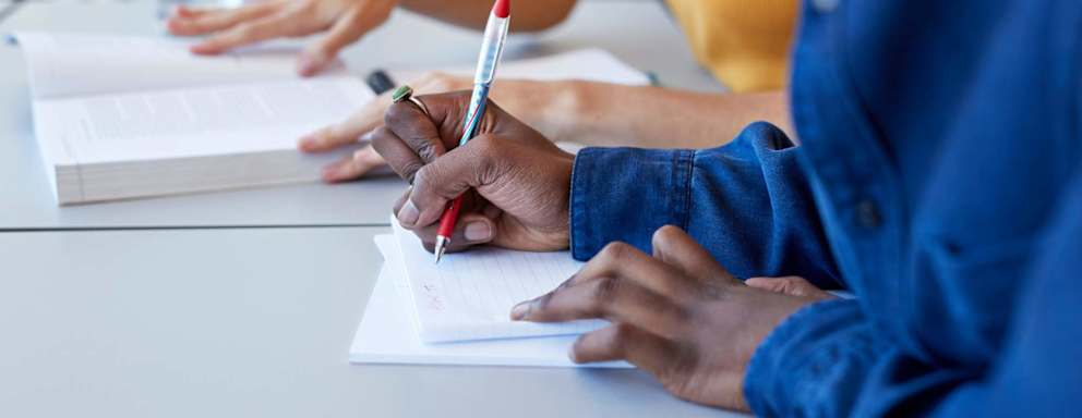 Close up of a Black male high school student writing in a notebook during a study session with a female friend. They are sitting at a table in a high school lunch room and looking at class textbook materials.