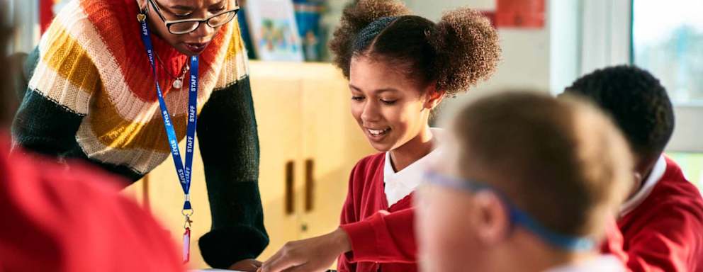 Cheerful elementary school girl showing her book to her teacher who is leaning over the desk and concentrating