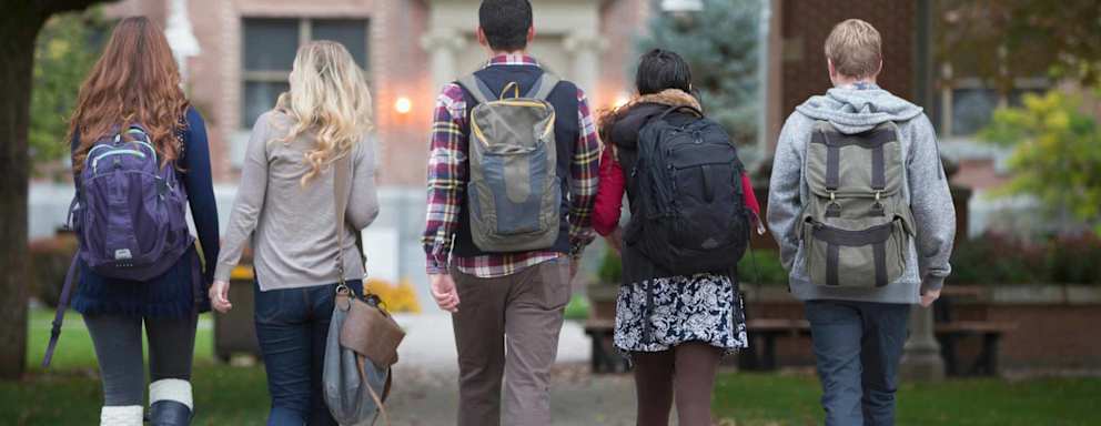 Group of college students walking together on a tree-lined campus path, carrying backpacks and chatting with each other.