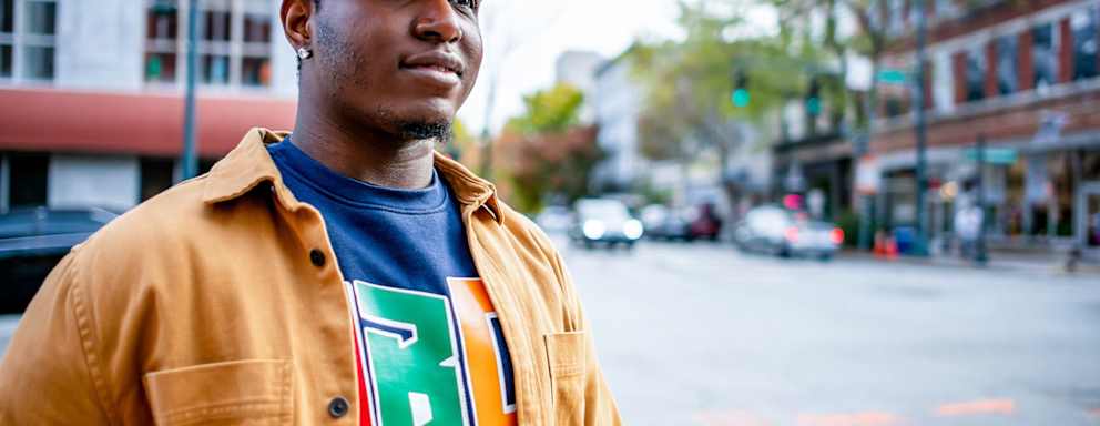 Black male HBCU college student wearing a cap and a blue sweatshirt with his school's HBCU logo walks downtown in a city on a fall afternoon.
