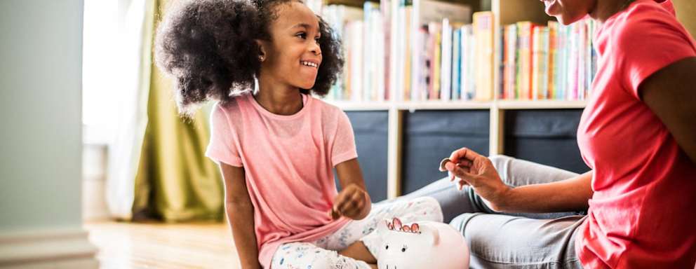 A Black mother and her young daughter are sitting on the floor in the living room at home. The mother is teaching her daughter about saving money as she helps count coins in her daughter's piggybank.