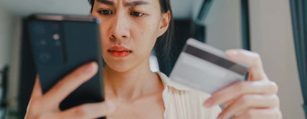 Asian American female college student using her credit card to pay her student loan bill. She is looking at her mobile phone, looking shocked at the amount she has to pay.