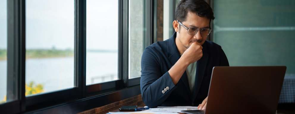 Asian American male Ph.D. student sitting at a table in a cafe. He is typing a report on his laptop computer. There are several paper notes on the table that he is referencing.