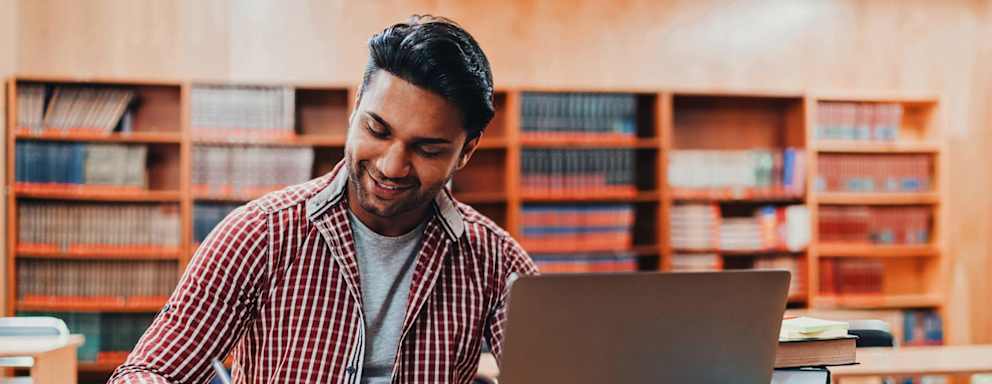 Male South Asian/Desi American college student studying in the library for his GRE exam. He has his laptop open on the library desk and is taking notes in a notebook.