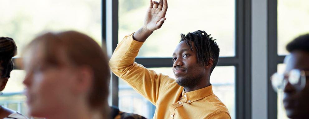 Black male community college student sitting at a desk in a classroom with other students. His arm is raised to answer a question from the professor off-screen.