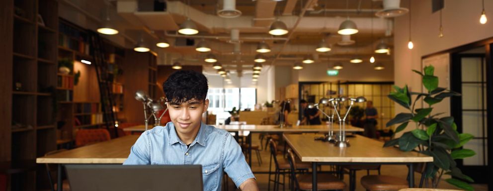 A male Asian American college student sitting at a table in a student center. He has his laptop open, and is using his credit card to pay for a school tuition bill online.