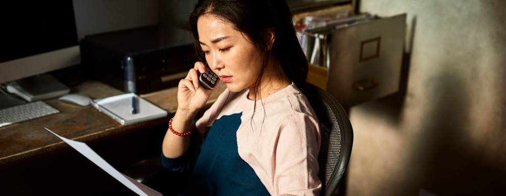 Serious mid adult Asian woman listening on phone, sitting in office chair in dark room, holding paperwork, checking accounts, financial planning, money concerns, difficulty, debt