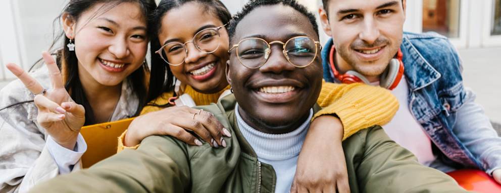A diverse group of college student friends after class. They are hugging each other and smiling while taking selfie photos.