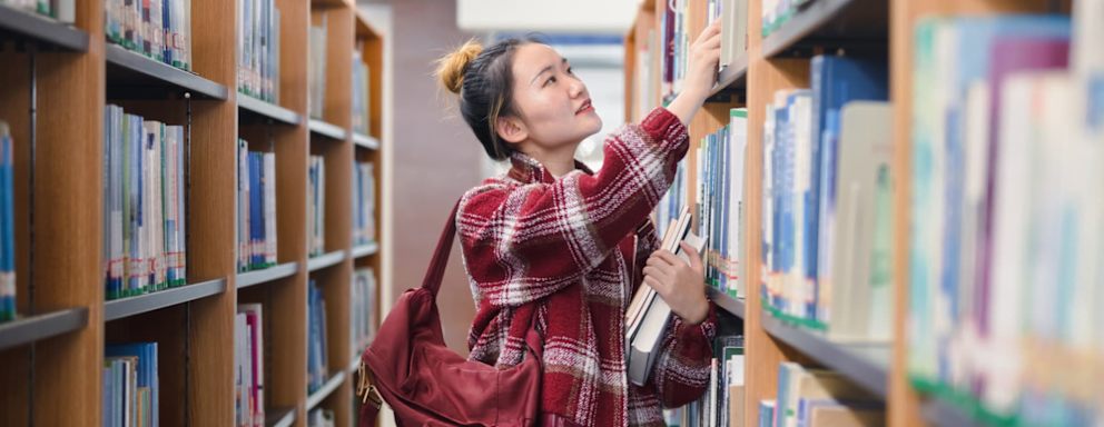 Female Asian American college student looking for textbooks on the shelves in her university's library.