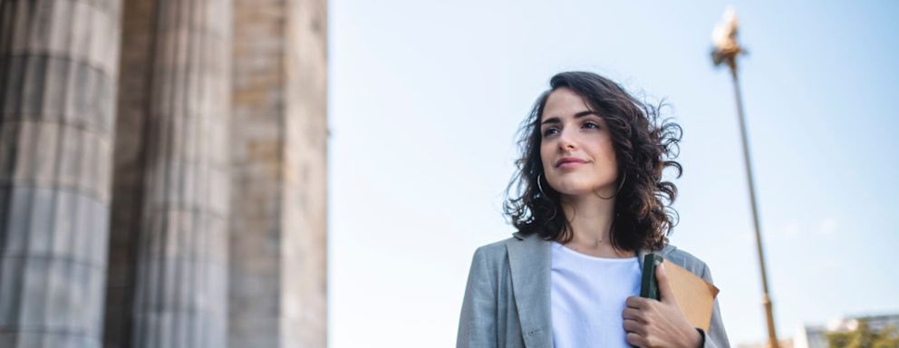 Female law student in her late 20s standing with a textbook at the entrance to a Neo-Classical university building on campus.