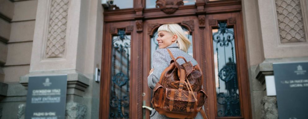 Female university student with blond hair walks towards a building entrance on campus with her backpack and smiles.