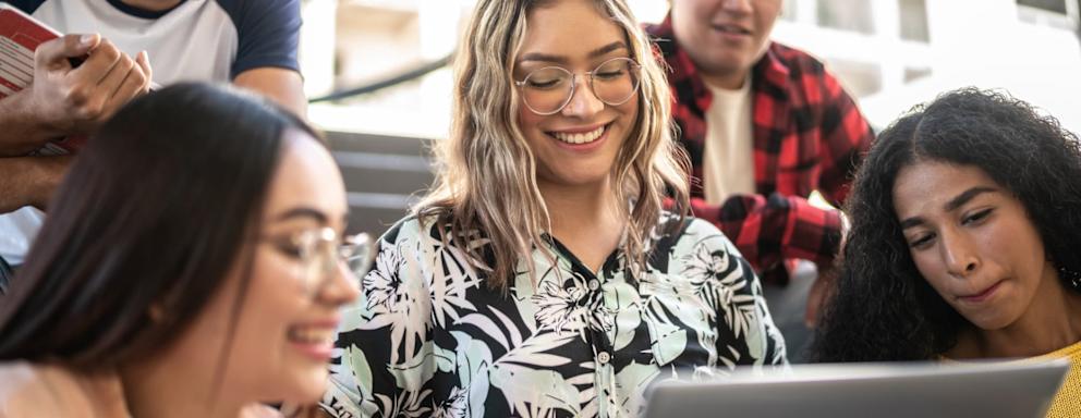 A Latina college student sits with her friends in a university classroom. She is smiling while showing them something on her laptop.