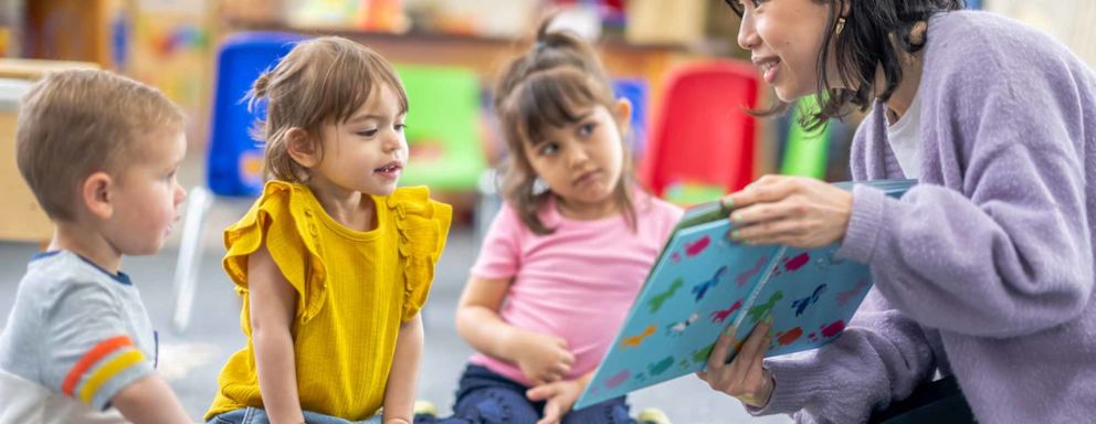 A preschool teacher sits on the floor of her classroom with a small group of students as she reads them a book.