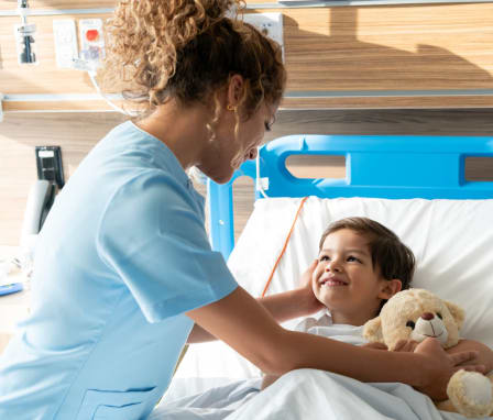 Nurse tucking in child patient in hospital bed with teddy bear