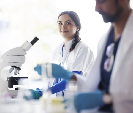 Two scientists working in a lab with a microscope wearing scrubs