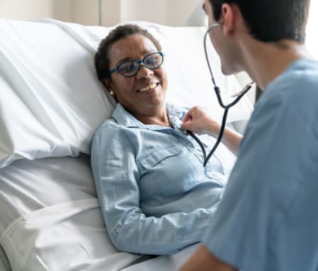 Female black patient lying down on hospital bed while nurse is checking her heart beat with stethoscope