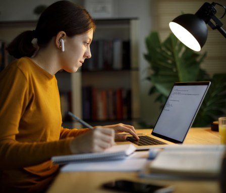 Woman typing on laptop and writing in a notebook under a desk lamp