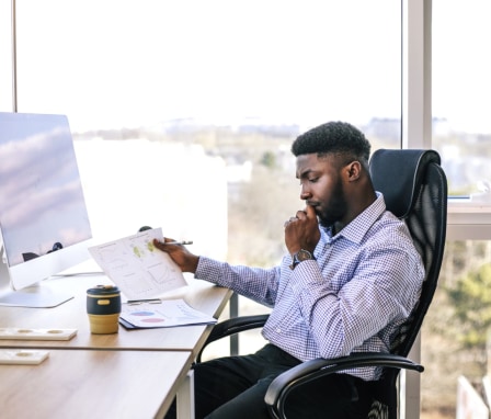Man looks at printed graphs in front of computer screen beside window