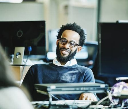 Man smiling at computer desk and talking to colleague