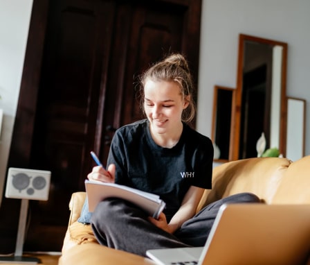 Woman taking notes in a notebook while looking at a laptop, while sitting on a couch