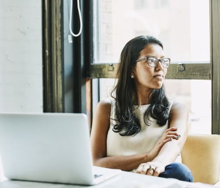 Woman looking out window, sitting at a desk with an open laptop