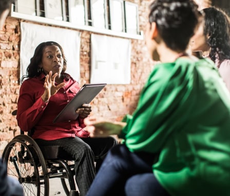 Woman in wheelchair holding tablet leading group discussion in office