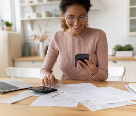 Smiling woman looking at phone with bills, a calculator, and a laptop in front of her