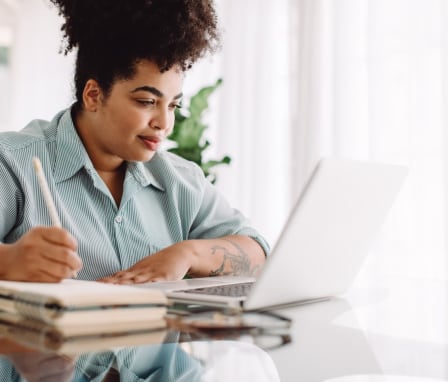 Woman taking notes in notebook while reading on laptop at home