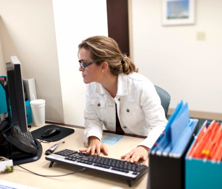 Mid-adult female administrative professional working at her desk in a nursing board office. She is looking at the computer screen while typing on the keyboard.