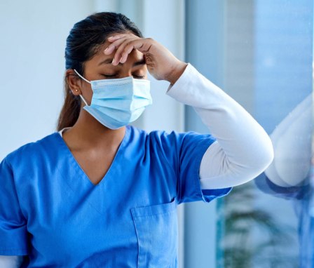 Young Asian-American female nurse with her eyes closed rubs her forehead while standing by a hospital window.