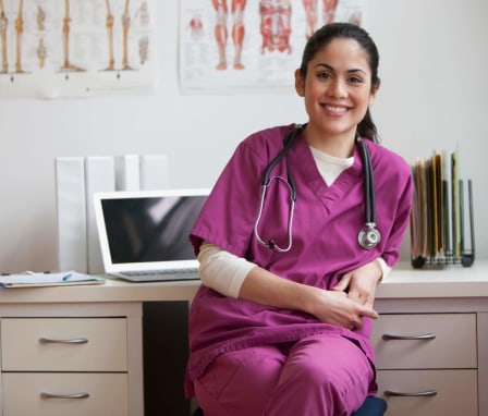 A female Hispanic nurse sitting at her desk in a clinical office. She is facing towards the camera and smiling. Her laptop and case files are on the desk behind her.