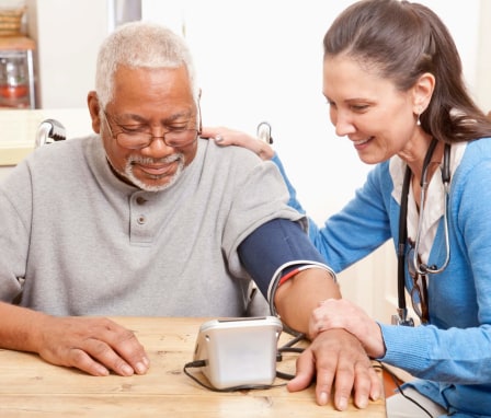 A mid-adult Caucasian female nurse is at an elderly Black patient's home taking his blood pressure. They are sitting at the patient's dining room table and smiling.