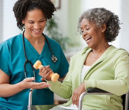 A mid-adult African-American home nurse is helping her elderly female patient in a wheelchair lift a hand weight. They are sitting together in the patient's home living room.