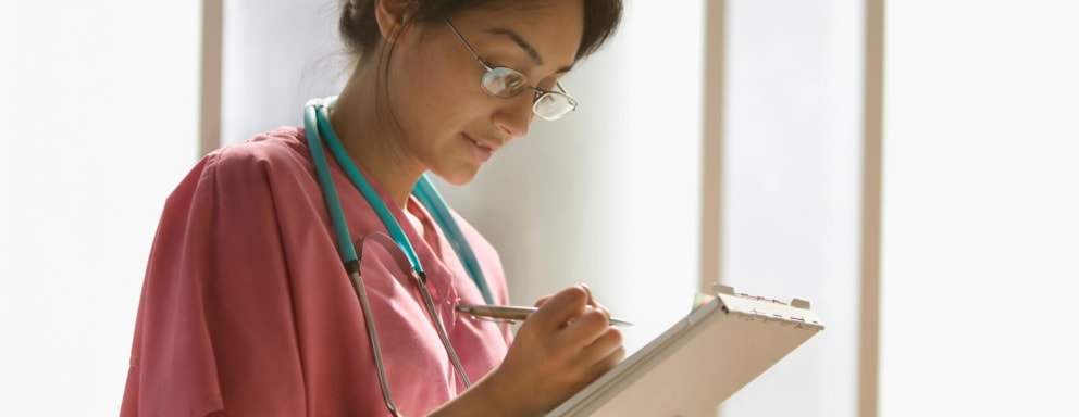A young Hispanic female nurse wearing eyeglasses and scrubs is writing notes on a clipboard.