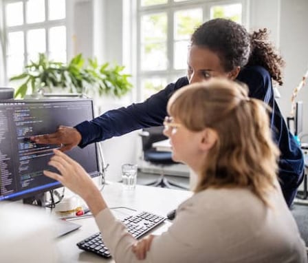 Lady pointing to a computer monitor showing her coworker how to troubleshoot code