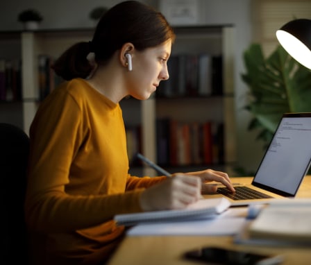 Woman typing on laptop and writing in a notebook under a desk lamp